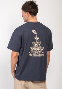 Muž v námořnickém tričku s grafikou šampaňského lahve, která se lije do naskládaných sklenic, a textem "Oslavujme" na zádech.