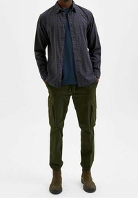 Camicia a scacchi con bottoni in blu scuro e verde, indossata sopra una maglietta blu navy. Pantaloni cargo verde oliva con tasche laterali. Stivaletti marroni alla caviglia.