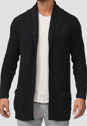 Hombre con cárdigan grueso de punto negro con dos bolsillos delanteros, camiseta blanca y pantalones gris claro, de pie frente a un fondo liso.