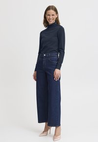 Maglione a collo alto navy con dettagli a bottone, abbinato a jeans scuri a gamba larga e tacchi nudi. Design semplice, aderente e casual.