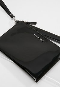 Pochette bracelet noire brillante Michael Kors avec fermeture éclair et bandoulière amovible sur une surface blanche.