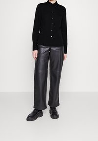 Camicia nera con colletto, abbinata a pantaloni in pelle nera a vita alta e gamba larga e scarpe nere chunky. Texture liscia in tutto.