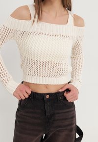 Pull en maille crème à épaules dénudées, avec un tissage ajouré et un ourlet côtelé. Associé à un pantalon en denim noir taille haute.