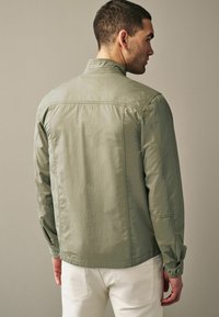 Veste légère vert clair avec un col montant, des poignets boutonnés et une texture lisse, vue de dos sur un fond neutre.