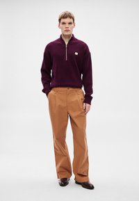 Bordó kötött pulóver félcipzárral és kis logóval, bézs bő szárú nadrággal és sötétbarna bőrcipővel párosítva.