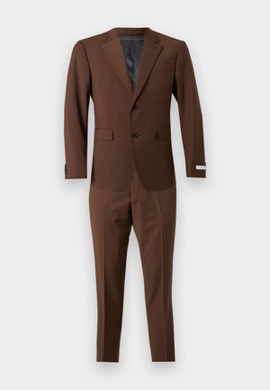 Mężczyzna stoi, ubrany w brązowy garnitur, jasnoniebieską koszulę z guzikami oraz brązowe eleganckie buty, na tle jednolitego jasnoszarego tła.