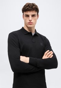 Jonge man met over elkaar geslagen armen, gekleed in een zwart poloshirt met lange mouwen, tegen een effen lichte achtergrond.