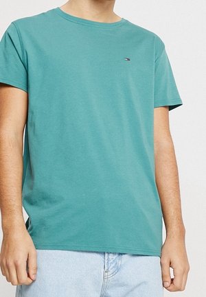 Man draagt een tealkleurig T-shirt met ronde hals en korte mouwen en lichtblauwe spijkerbroek, staand tegen een effen lichte achtergrond.