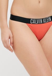 Röda ribbade stringtrosor med svart elastiskt midjeband som har texten "CALVIN KLEIN" i vitt, vilket framhäver deras sportiga design.