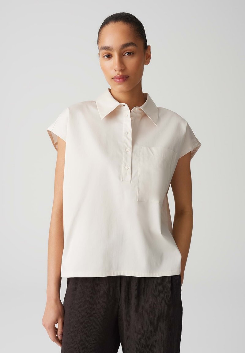 Lichtbeige blouse met korte mouwen, een kraag, knoopsluiting en een enkele borstzak; gladde stof, ontspannen pasvorm, minimalistisch ontwerp.