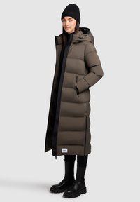 Vrouw in een lange olijfgroene pufferjas met capuchon, zwarte beanie, zwarte leggings en chunky zwarte laarzen, staand met de handen in de jaszakken.