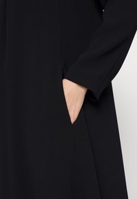 Robe noire à la texture lisse, dotée d'une poche latérale et de manches longues, montrant une main reposant sur le bord de la poche.