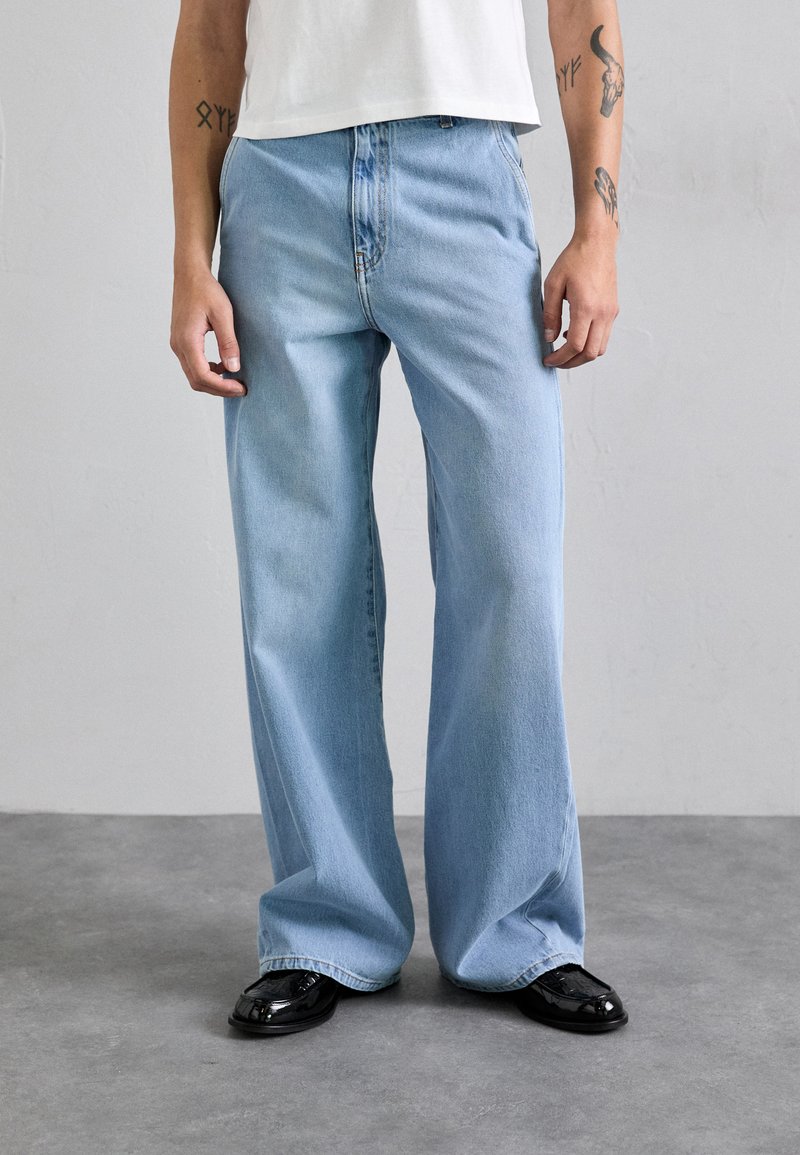 Svetlo modri široki jeans iz denima, z značilnim oblikovanjem petih žepov in zadrgo. Oblečen z belo majico in črnimi čevlji.