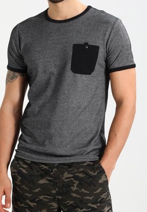 Mann trägt graues T-Shirt mit kurzen Ärmeln, schwarzer kontrastierender Tasche und Besatz, kombiniert mit Tarnshorts, eine Hand in der Tasche.