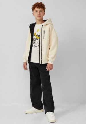 Crème-kleurige fleece jas met een zwarte capuchon, ritssluiting aan de voorkant en een wit T-shirt met grafisch ontwerp eronder, gecombineerd met zwarte broek en witte sneakers.