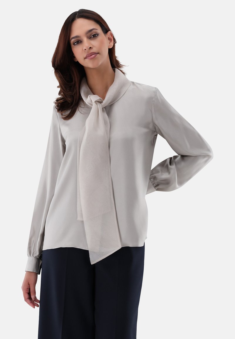 Blusa grigia chiara con una sciarpa legata al collo, tessuto morbido, maniche lunghe con polsini a bottone e un fit rilassato.