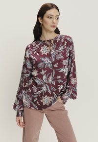 Langärmlige, burgunderfarbene Bluse mit floralem Muster in Blau und Creme. Verfügt über einen gebundenen Ausschnitt und weite Ärmel. Glatte Stofftextur.