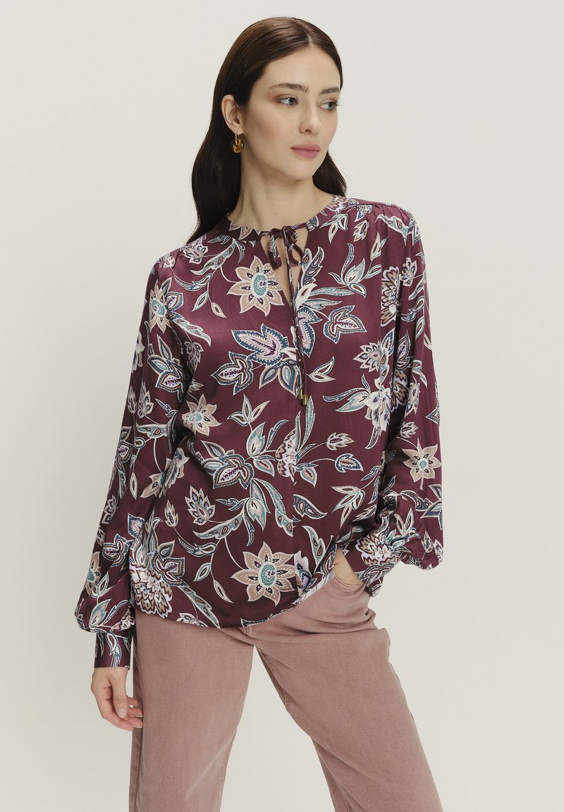 Langärmlige, burgunderfarbene Bluse mit floralem Muster in Blau und Creme. Verfügt über einen gebundenen Ausschnitt und weite Ärmel. Glatte Stofftextur.