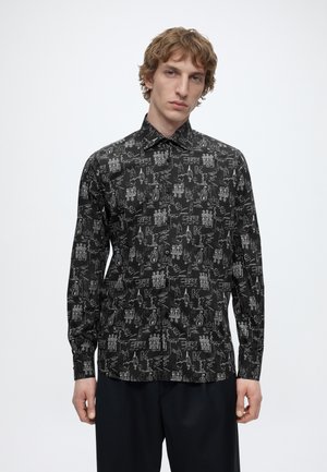 Jeune homme portant une chemise noire à boutons avec des croquis abstraits blancs et du texte, debout devant un fond blanc uni.