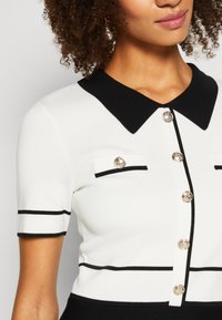 Femme portant un haut blanc à manches courtes avec col noir, bordure noire, boutons décoratifs dorés et rabats de poche sur fond uni.