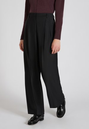 Pantalons plissés noirs en tissu lisse, à taille haute, avec poches latérales et coupe décontractée, associés à des mocassins noirs.