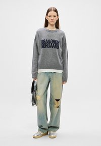 Suéter de malha cinza com letras em azul-marinho, corte solto. Usado com jeans claros e rasgados e ténis prateados. Bolsa refletora na mão.