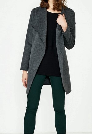 Femme portant un manteau ouvert gris foncé sur un haut noir et un pantalon slim-fit vert foncé, se tenant contre un fond blanc.