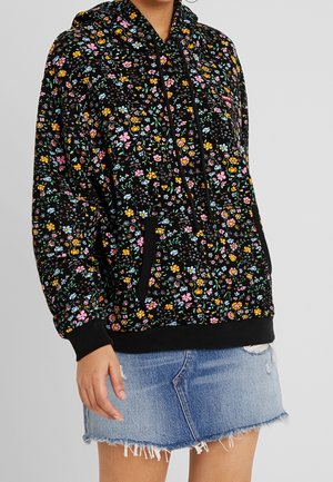 Person trägt einen schwarzen Kapuzenpullover mit bunten kleinen Blumenmustern und schwarzen Bündchen, kombiniert mit einem hellen Jeansrock mit ausgefranstem Saum.