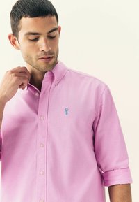Camicia button-up rosa chiaro con colletto ripiegato, caratterizzata da un piccolo logo ricamato blu e dettagli con bottoni sul frontale.