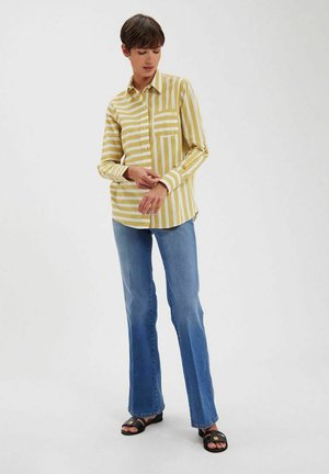 Chemise à rayures jaunes et blanches à manches longues avec col et poche avant, associée à un jean évasé bleu clair et des sandales noires.