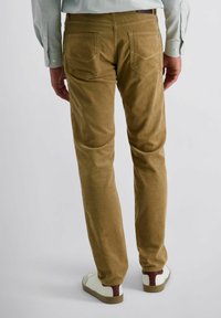 Homme portant un pantalon en velours côtelé marron, des baskets blanches avec des accents bordeaux, et une chemise à manches longues bleu clair, se tenant devant un fond uni.