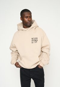 Sudadera color beige hecha de un material suave con un bolsillo frontal. Presenta texto impreso en negro "MULTIPLY ORIGINALS EST 2015" en el pecho.