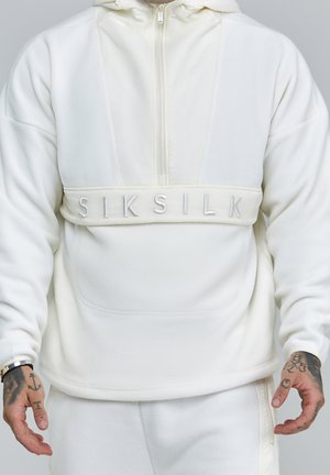 Mężczyzna noszący biały polarowy kaptur z półzamkiem i haftowanym napisem „SIKSILK” na piersi, pokazujący tatuaże na rękach i zegarek na nadgarstku.