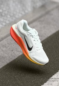 Παπούτσι τρεξίματος Nike με λευκό διχτυωτό επάνω μέρος, μαύρο λογότυπο swoosh και πολύχρωμη σόλα με γκριάντ από πορτοκαλί σε κίτρινο.