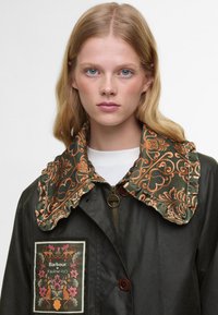 Casaco verde com um colarinho floral laranja bordado, fecho de zíper e um patch decorativo. Feito de um material liso e texturizado.