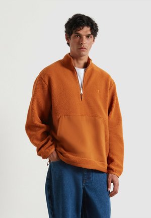 Pullover in pile arancione con chiusura a metà zip e tasca a toppa. Vestibilità oversize, materiale tessurizzato e polsini a costine. Abbinato a jeans blu.