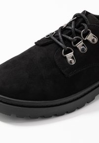 Chaussure en daim noir avec un bout rond, surface texturée, trois œillets en métal et lacets noirs ; dotée d'une semelle épaisse et robuste noire.