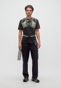 Νέος άνδρας με μαύρη εξωτερική ενδυμασία και πράσινο λουρί στήθους, κρατώντας γκρι παγούρι νερού, στέκεται μπροστά σε μονόχρωμο φόντο.