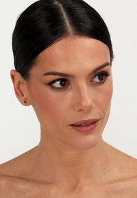 Gros plan du visage d'une femme, avec une peau lisse, des cheveux noirs en chignon lisse, des boucles d'oreilles en stud avec une petite pierre précieuse verte, un maquillage neutre.