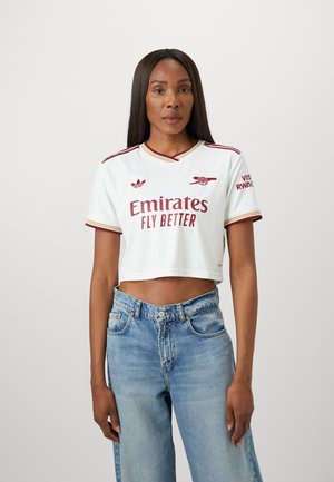 Kvinna med långt rakt hår iförd en vit kortärmad Arsenal fotbollströja och blå högmidjade jeans mot en enfärgad bakgrund.