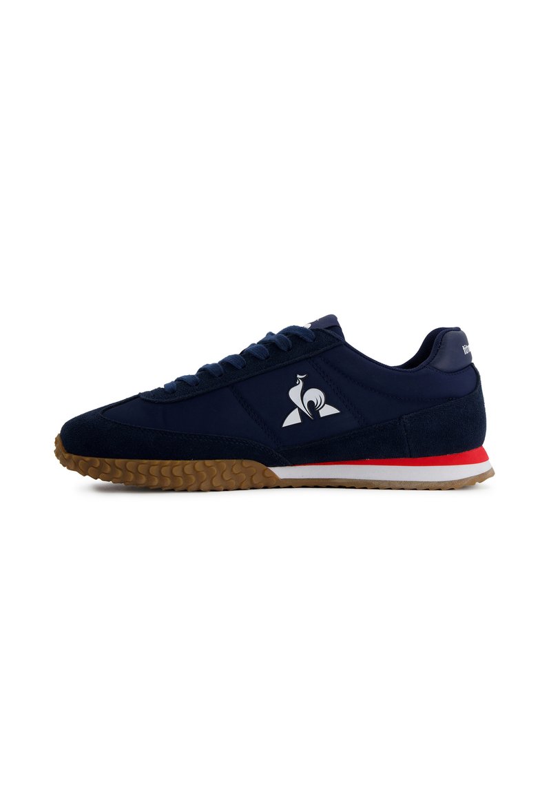 Baskets bleu marine avec semelle texturée marron, rayure rouge et blanche au milieu, logo de coq blanc sur le côté et lacets assortis bleu marine.