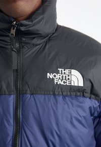 Pufferjakke med en sort overdel og en marineblå nederdel, med en høj krave og detaljeret lynlås. The North Face-logoet er tydeligt vist.