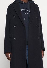 Cappotto doppiopetto di un blu scuro in misto lana, con revers a punta e grandi bottoni, abbinato a una felpa blu navy sotto.