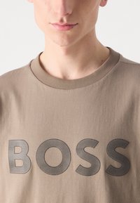 Šviesiai rudas trumpas rankovės marškinėliai su apvaliu kaklu, su dideliu, tekstūriniu juodu "BOSS" logotipu krūtinėje. Pagaminta iš minkštos medvilnės medžiagos.