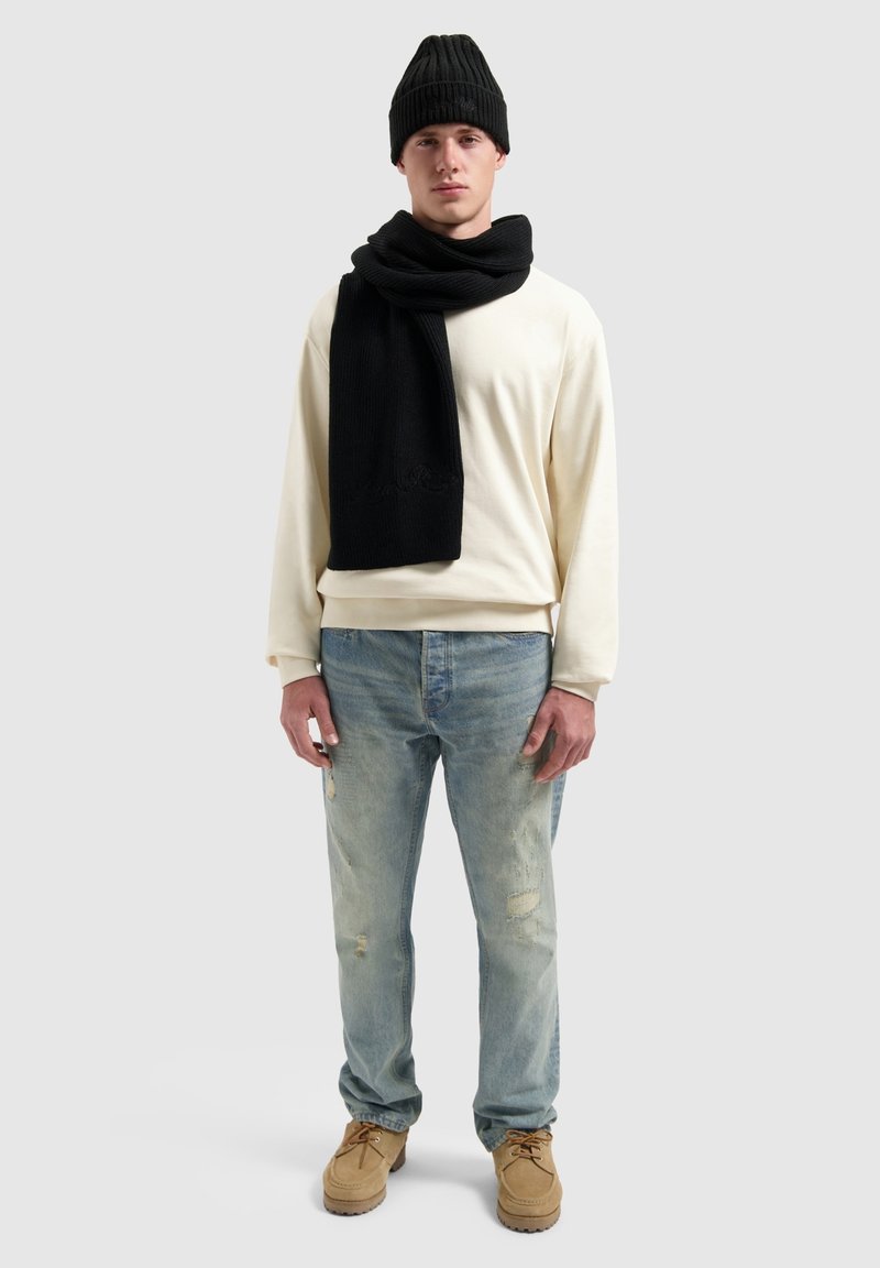 Creme sweatshirt, zwarte gebreide sjaal en beanie; lichtblauwe versleten jeans, tan schoenen met veters, staand tegen een grijze achtergrond.