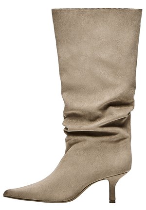 Botte en daim beige montant au genou avec bout pointu, tige souple et talon bas et fin, vue de côté sur fond blanc.