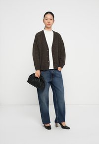 Dunkelbraune Strickjacke mit einer lockeren Passform, ausgestattet mit sechs Knöpfen. Getragen über einem weißen Hemd, kombiniert mit blauen weiten Jeans und schwarzen Heels.