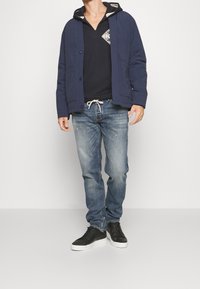 Mörkblå quiltad jacka över en svart hoodie, ljusa denimjeans och svarta sneakers. Funktioner inkluderar knäppning och dragsko i midjan.