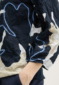 Haut à motifs bleu et blanc avec une texture froissée, présentant des poignets élastiqués et une coupe ample. Accents bleu marine le long du design.
