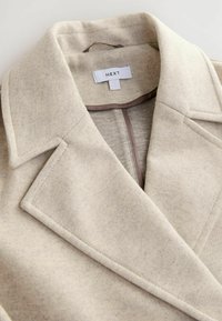 Abrigo de mezcla de lana beige con cuello de muesca, abertura frontal y un diseño minimalista. La etiqueta dice "NEXT" con un acento gris en el interior.